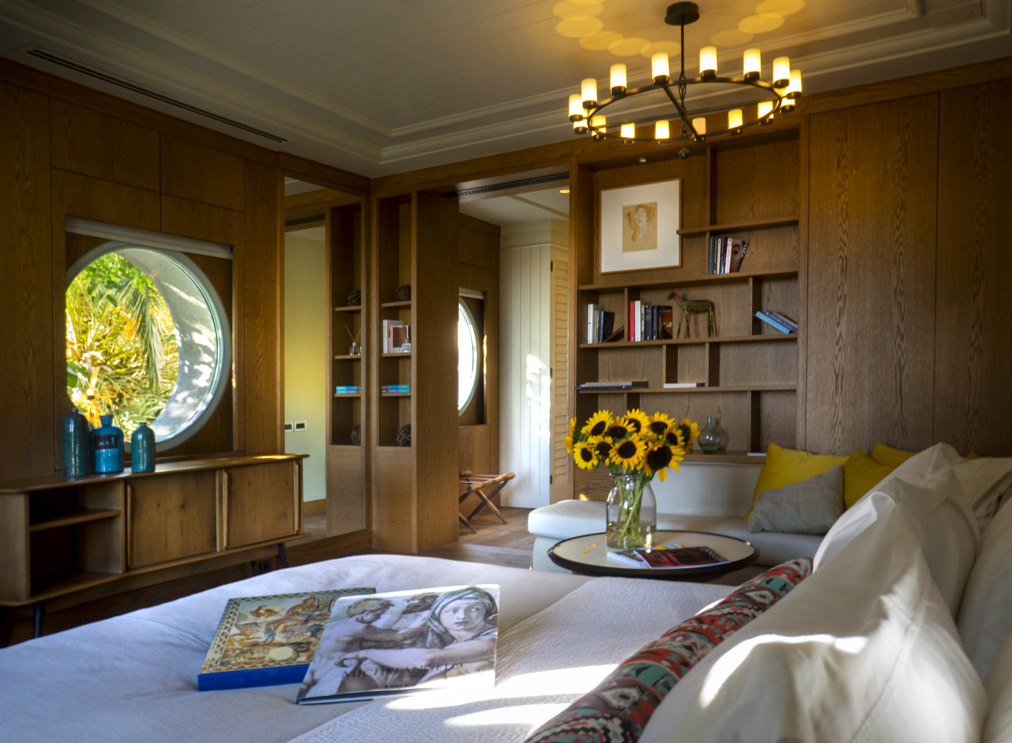 A bedroom lined in blond wood with a round window and a framed still life