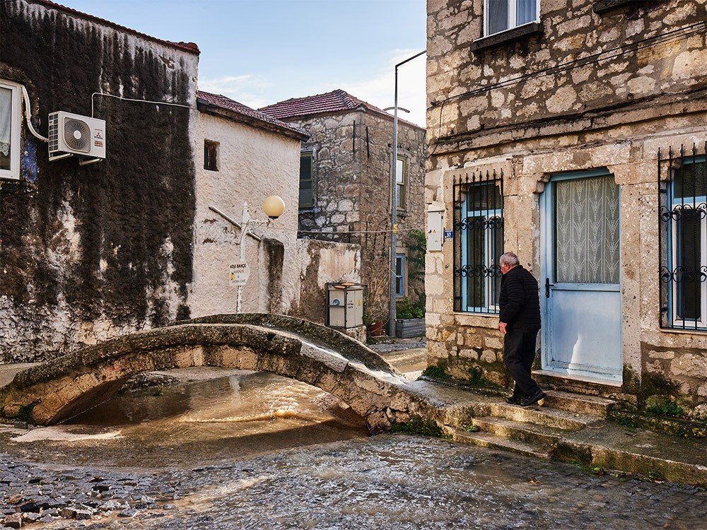 A quiet Alaçatı lane of old stone houses and a low bridge