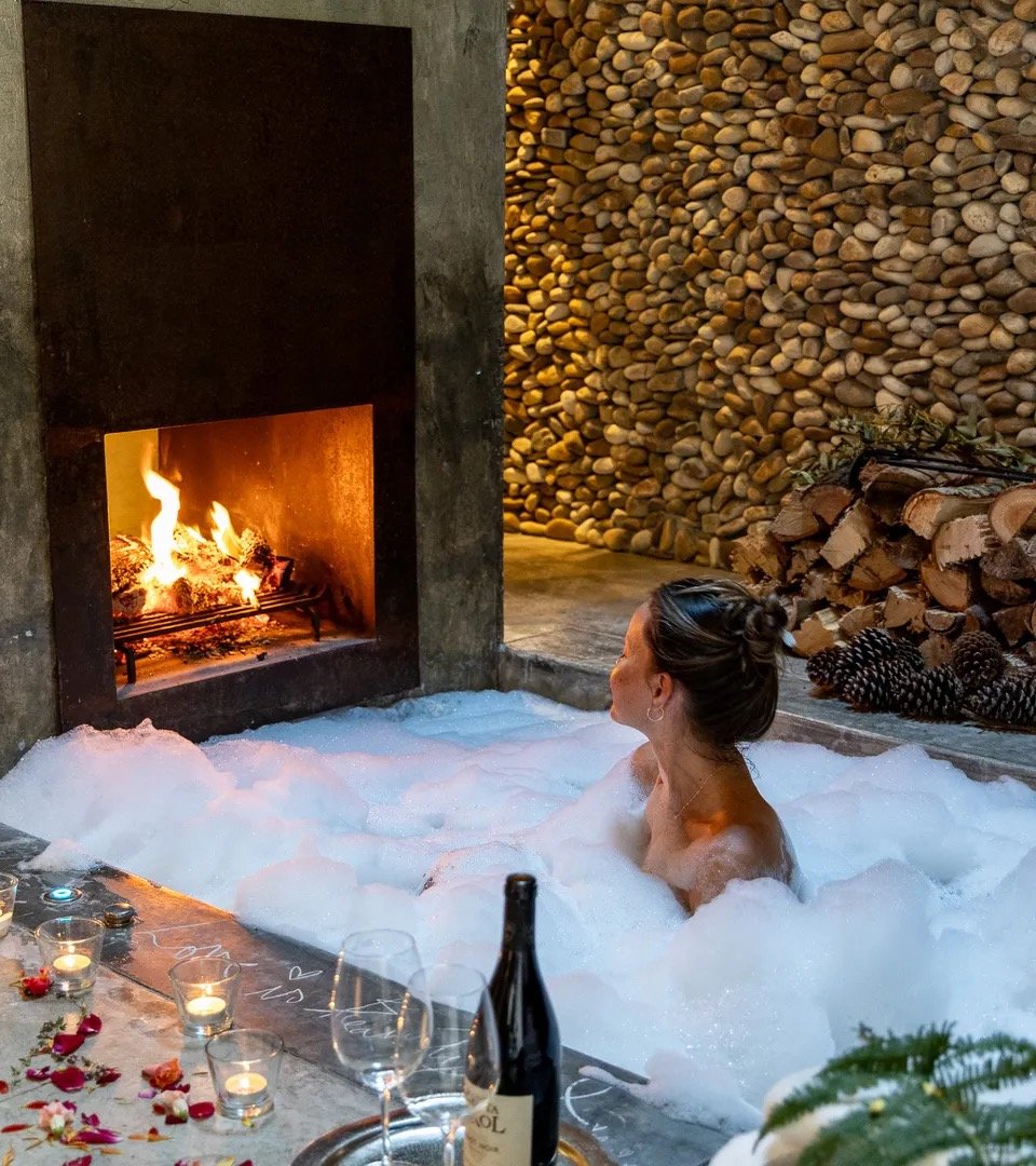 An outdoor stone bath beside an open fire, foam and candlelight against a pebble wall