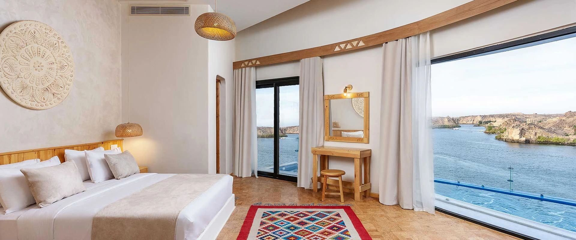 A room at BenBen by Dhara, white plaster walls, a carved Nubian medallion above the bed and a full wall of glass onto the Nile and the far bank