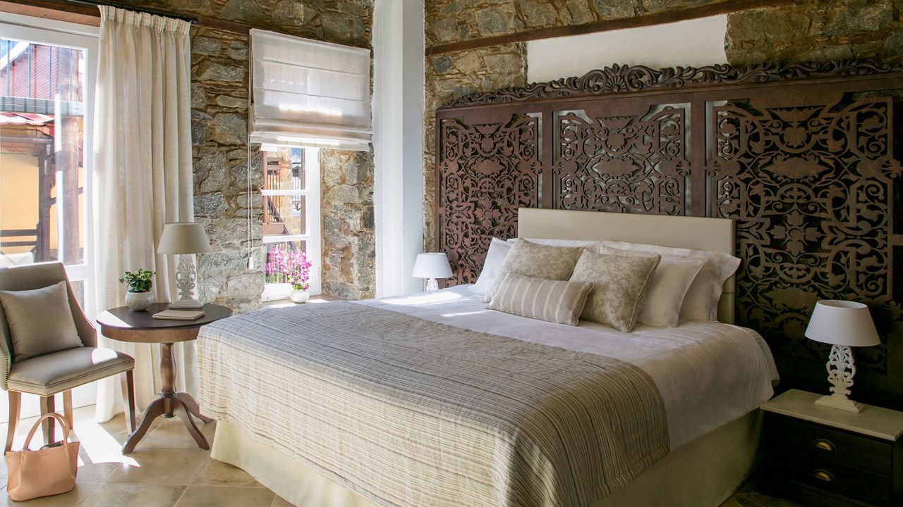 A bedroom with bare stone walls, a carved wooden headboard and village views
