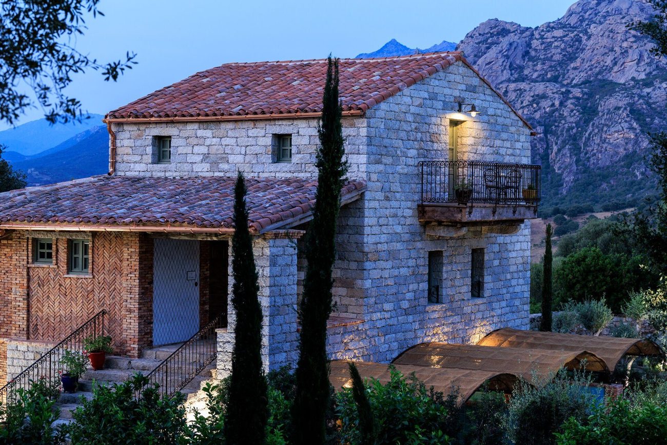 A restored granite shepherd's house at dusk, a cypress beside it and the mountains behind
