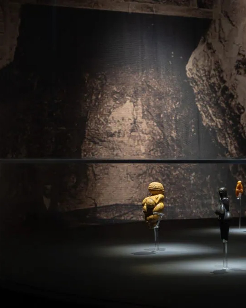 Gold and dark replicas of Paleolithic Venus figurines staged against a large cave photograph