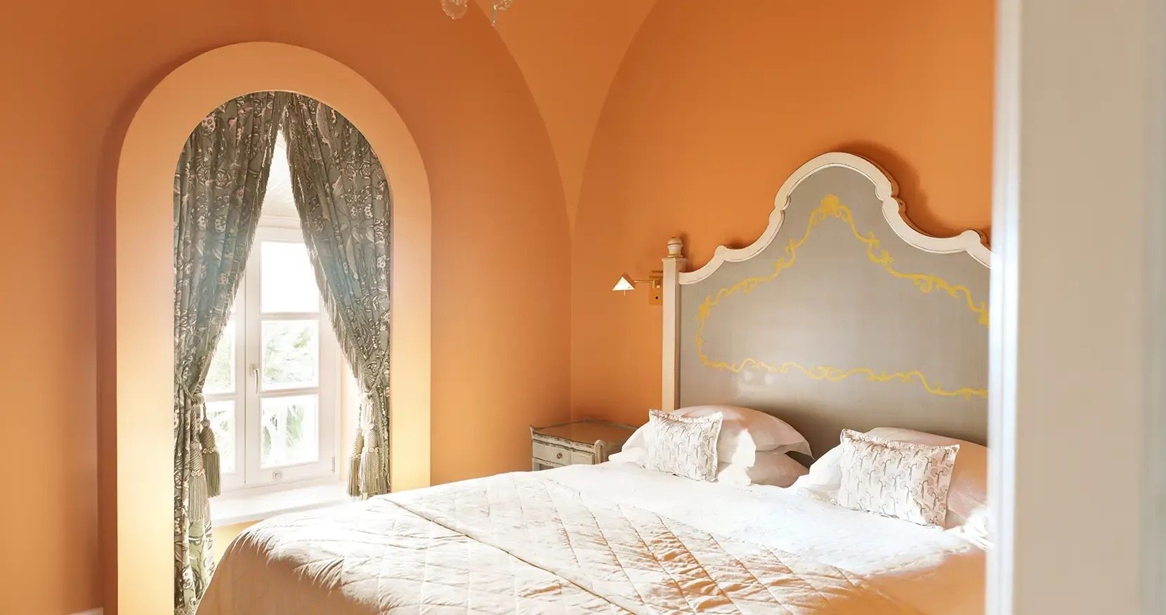 A vaulted suite in apricot, with a painted Venetian headboard and an arched, curtained window
