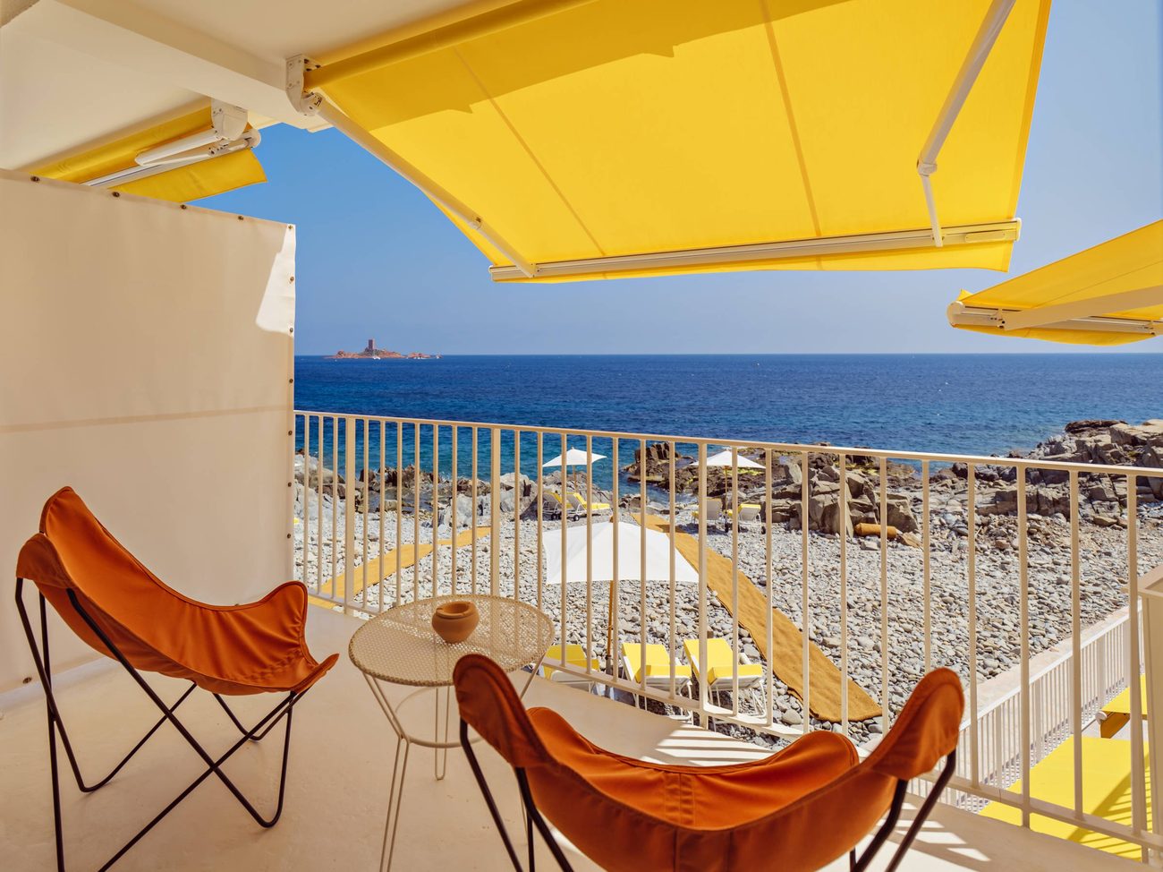 A sea-view room terrace with orange butterfly chairs under a yellow awning above the rocks
