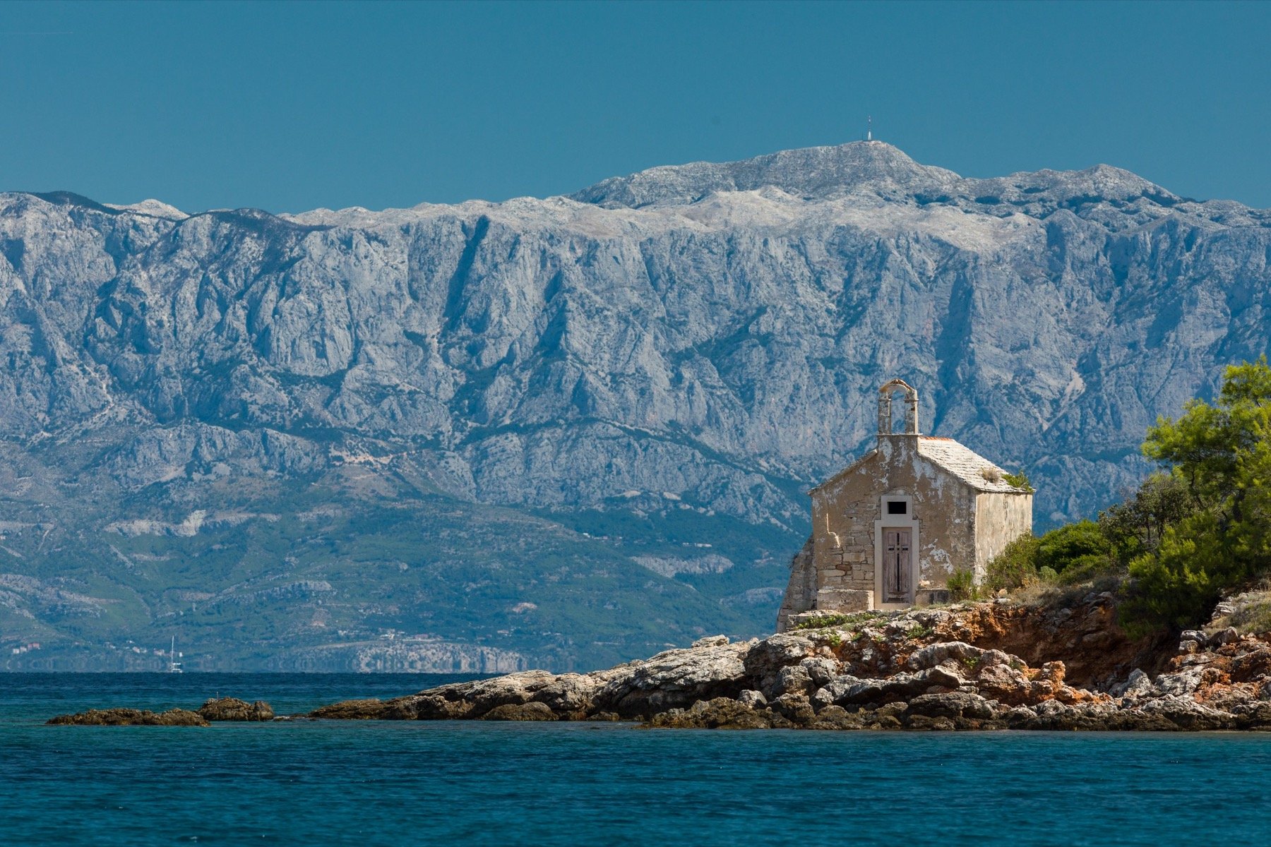 A stone chapel on a rocky point with the Biokovo mountains across the channel