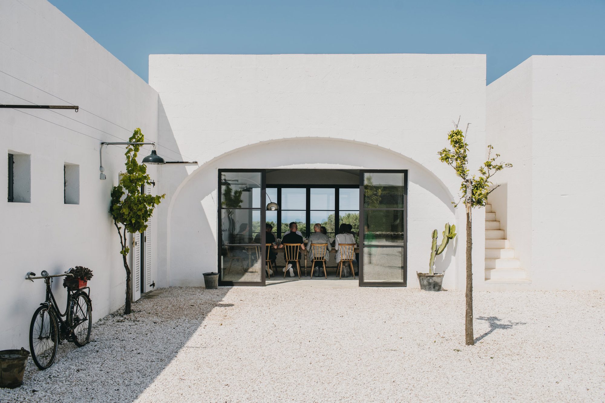 The arched glass doorway of Masseria Moroseta opening off the gravel courtyard, with guests at the long table inside, a vine and a bicycle against the white wall