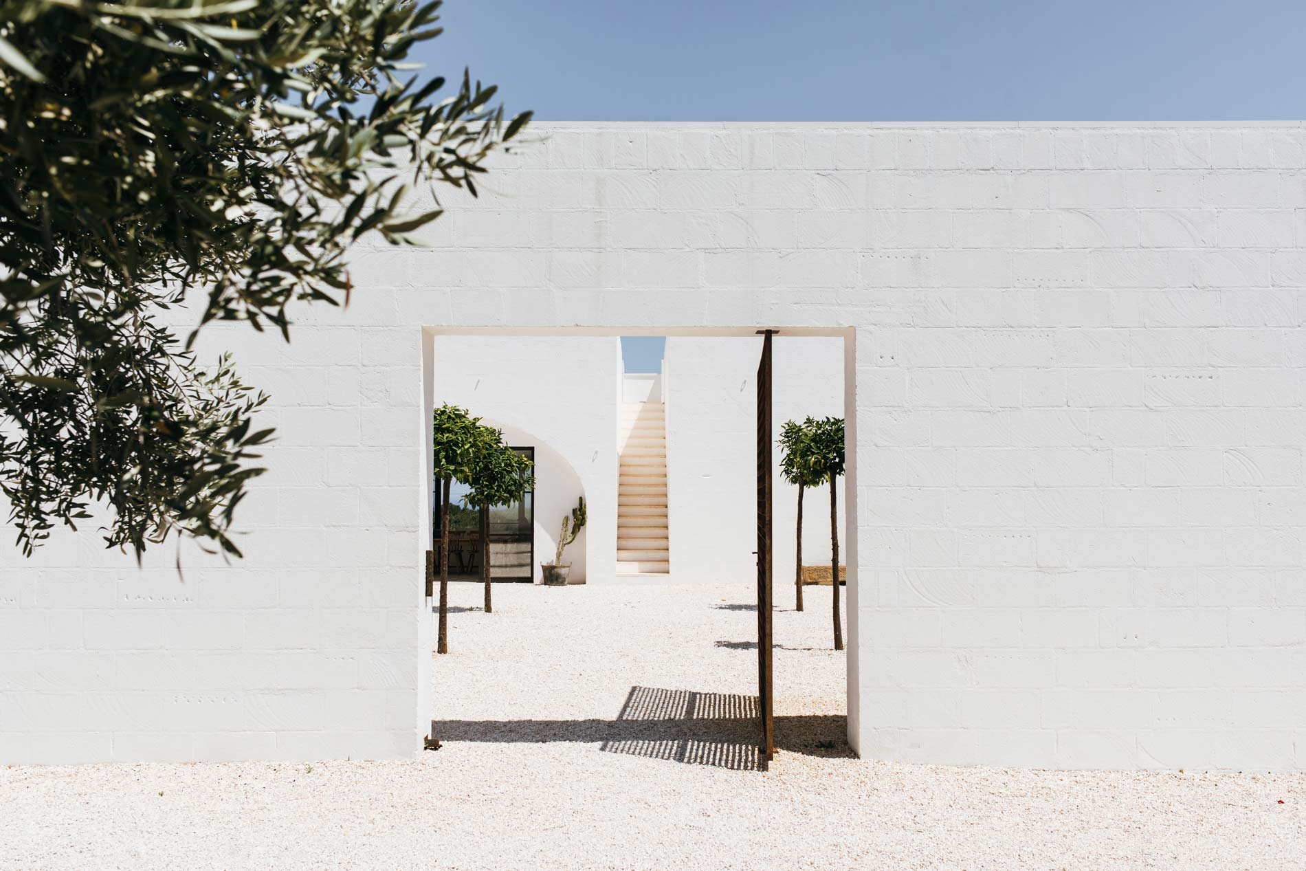 A doorway cut through the white perimeter wall of Masseria Moroseta, framing the gravel courtyard, potted lemon trees and a pale stone stair beyond