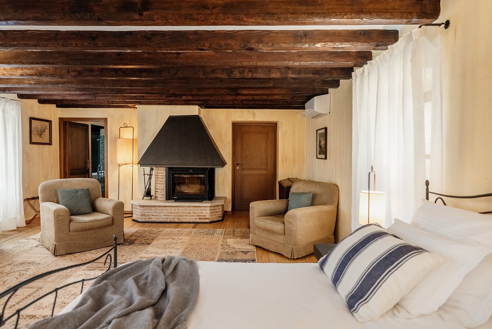 A rustic suite under dark beams with an open fireplace and an iron bed