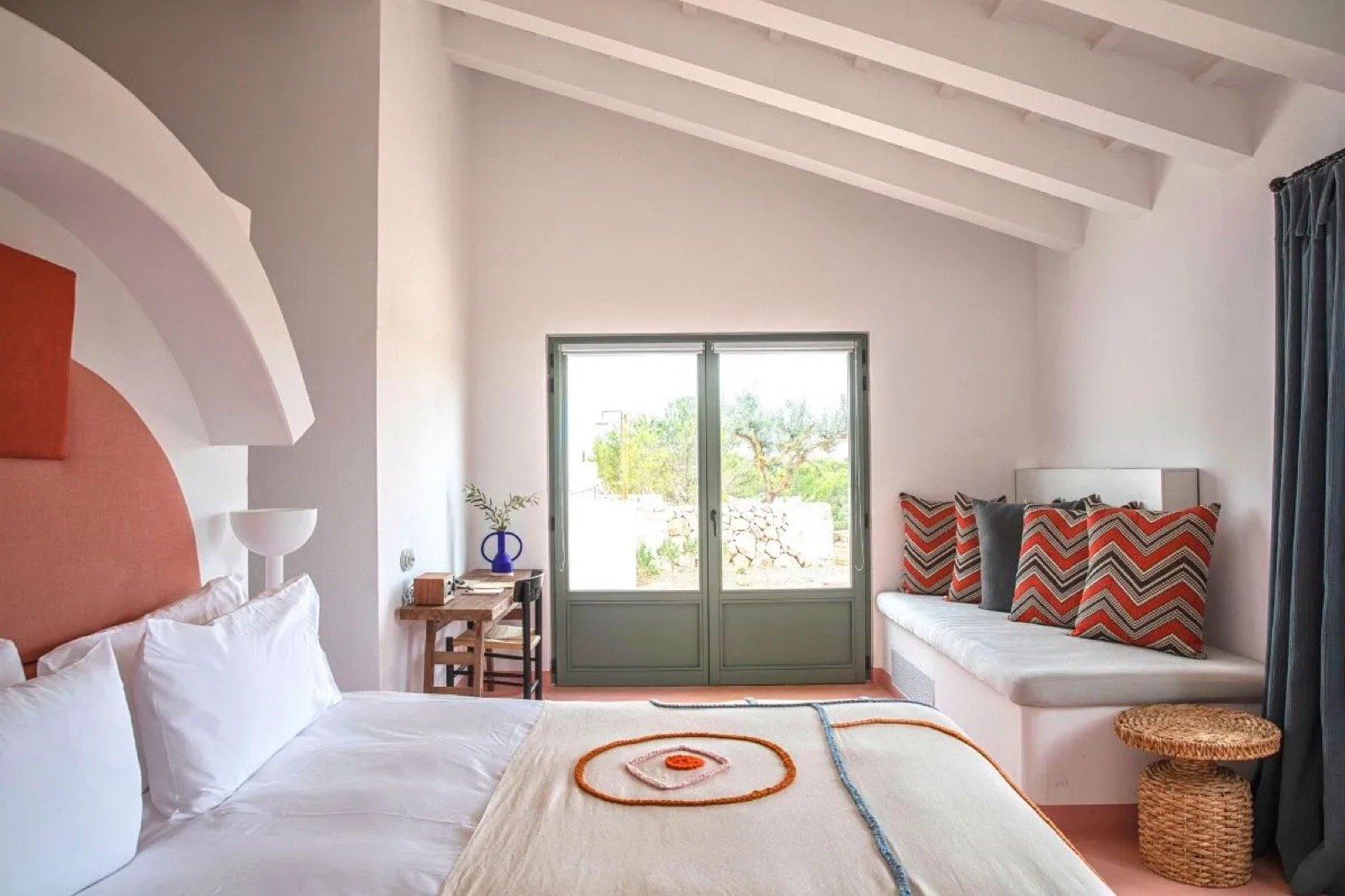 A guest room with a coral arched headboard, pale plaster and green shuttered doors to the garden