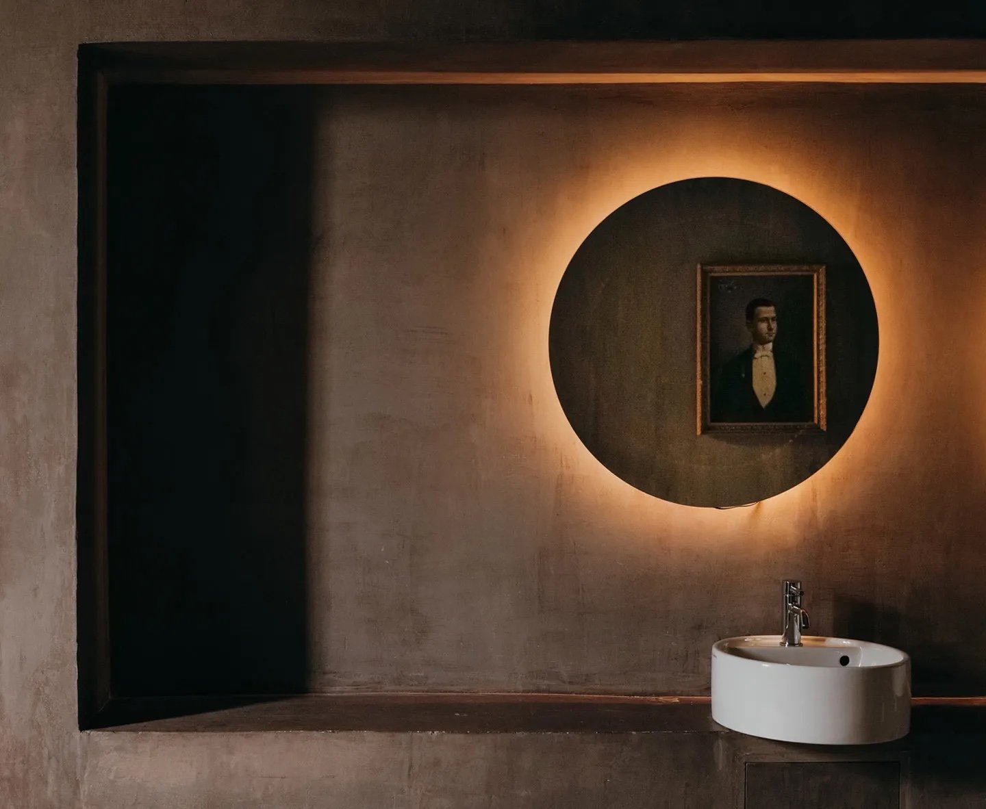 An ash-grey bathroom with a backlit round mirror and an antique portrait