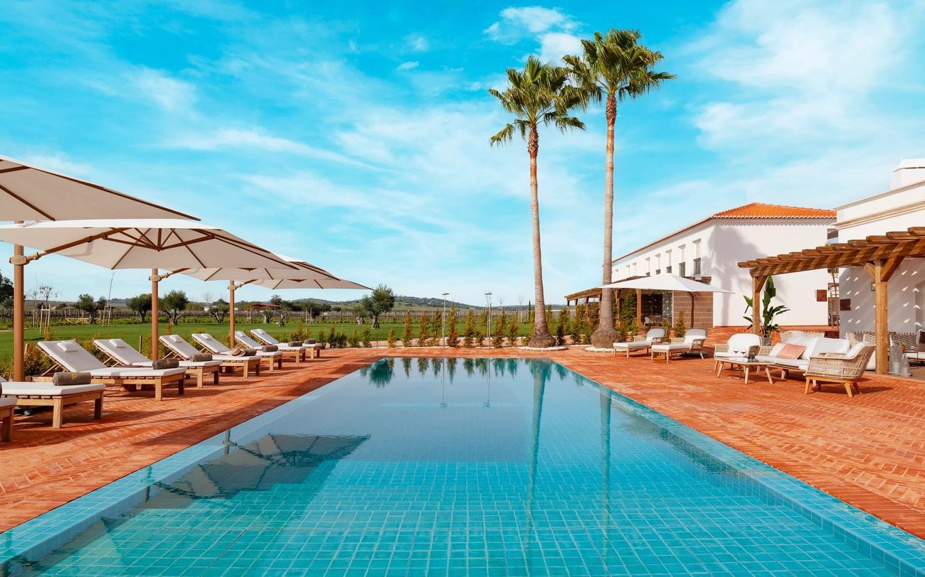 The outdoor pool with loungers, palms and the estate beyond under an Alentejo sky