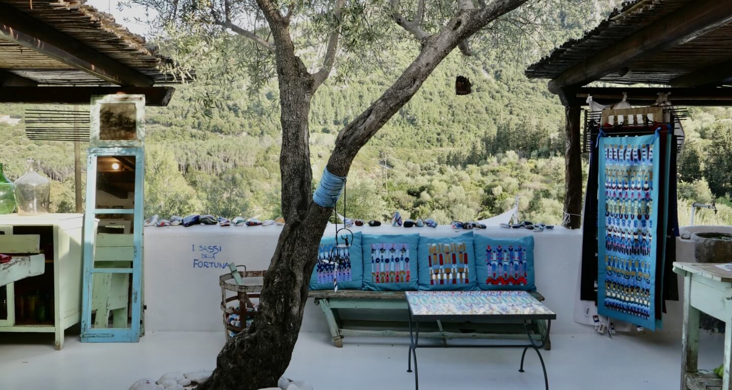 A craft terrace hung with Sardinian textiles and jewellery under an olive tree