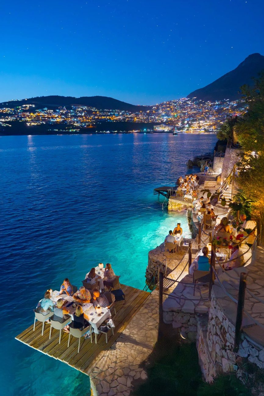 Lamplit tables on the waterfront terrace at night, the lights of Kalkan across the bay
